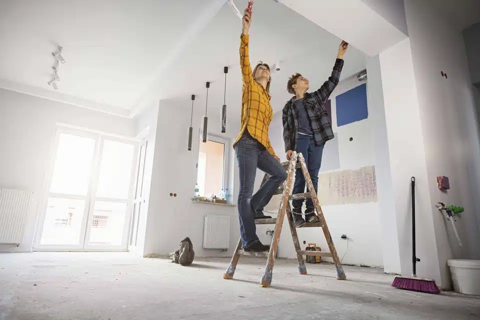 Two people doing home renovations standing on a ladder preparing walls to paint.