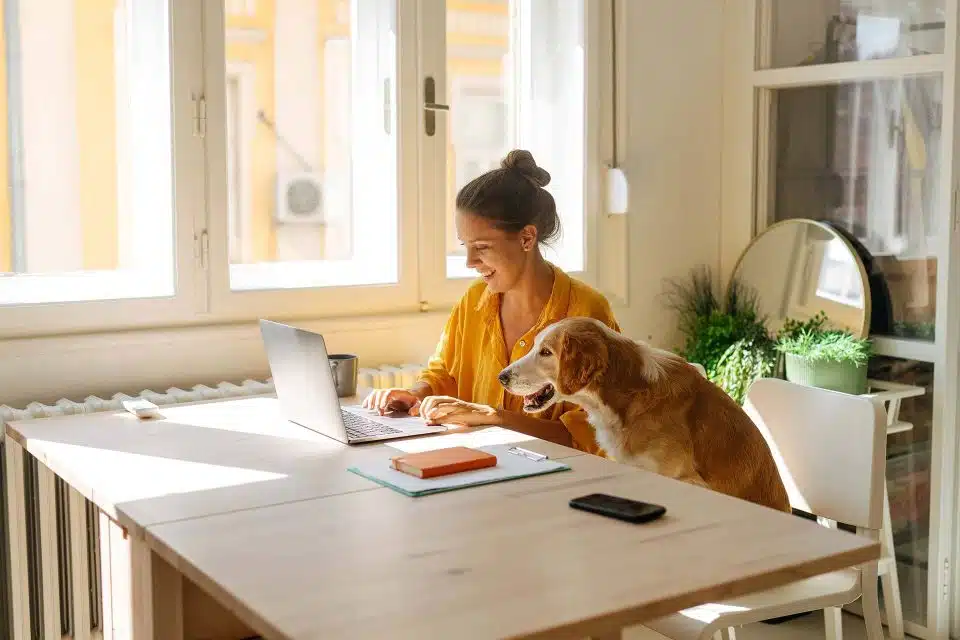 Woman with brown hair in a bun smiling at her computer as she searches for a home to buy with her dog sitting on the chair next to her