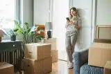 blond woman in striped shirt smiling and looking at her phone after buying her first home surrounded by moving boxes